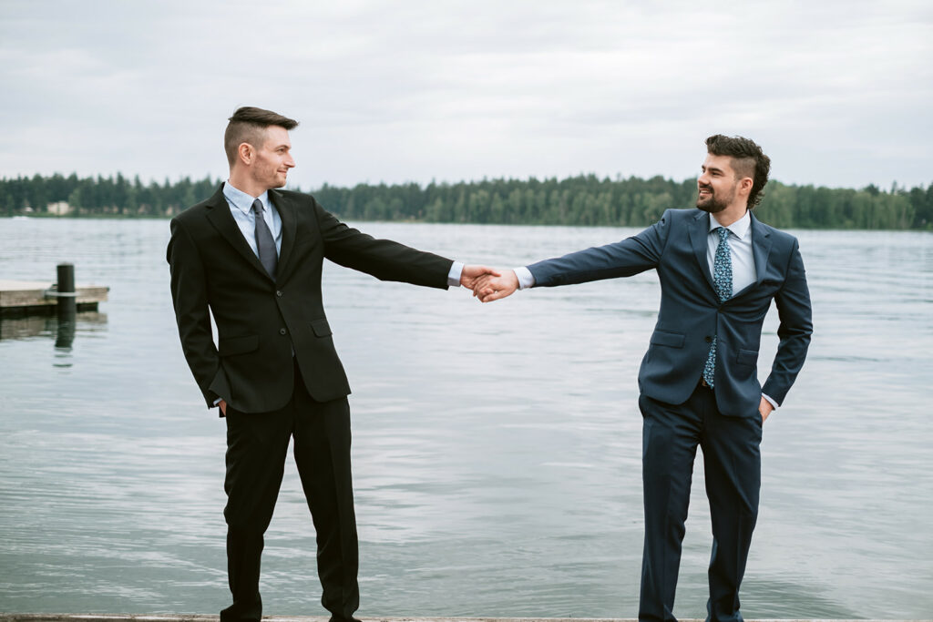 two grooms hold hands in front of lake