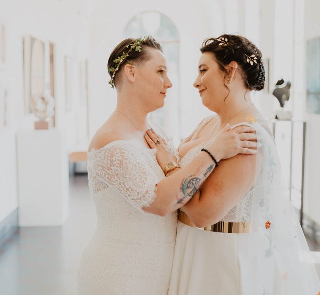 Juliana and Nicole smiling at each other wearing white dresses on their wedding day